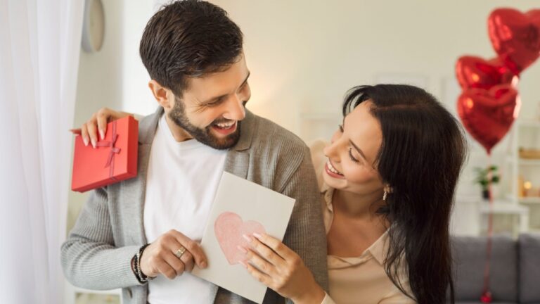 Portrait of a young pretty smiling woman holding greeting card and red present box and hugging her boyfriend. Happy couple in love. Care, congratulations, tenderness and Valentines day concept.