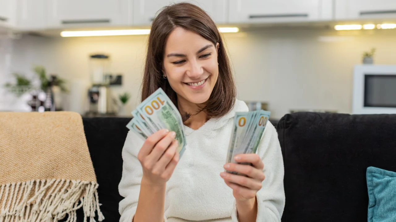 Young woman at home counts dollar cash smiles at total plans invest and plans dream purchase. Caucasian girl on sofa holds money stack checks sum again feels joy and calm and hopes for bright future