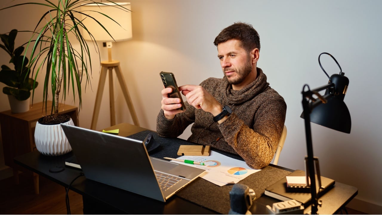 Man sitting at desk using smartphone while working in a home office with laptop and documents. Tired freelancer procrastinating at workplace during remote work. Unproductive man using phone