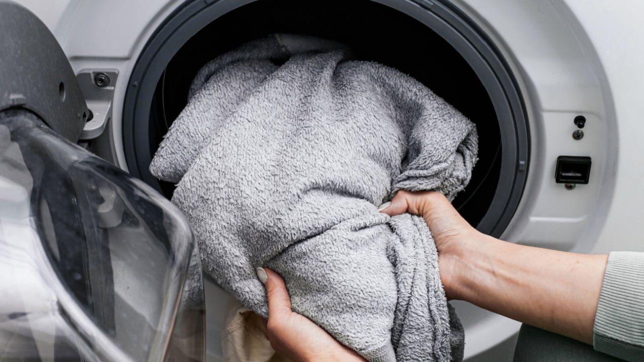Washing blanket in front load washing machine. Hands loading soft fabric textile. Proper laundry temperature care. Home washing process detail. Fabric cleaning routine. Household appliance use.