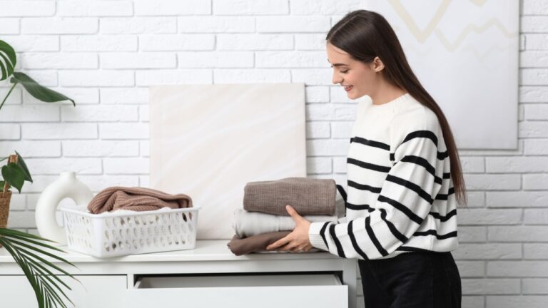 Young woman putting stack of warm sweaters into drawer at home