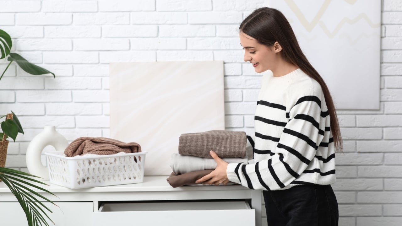 Young woman putting stack of warm sweaters into drawer at home