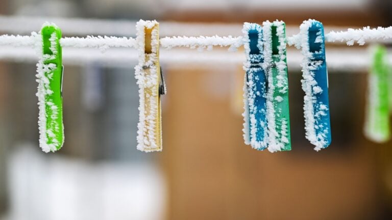 A line of clothespins are covered in snow. The clothespins are green and blue. The snow is covering the clothespins and the clothesline