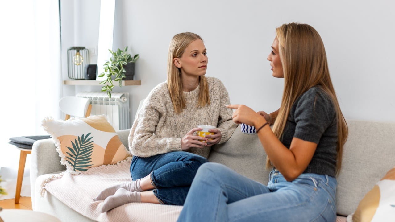 Two young women having a serious conversation while sitting on a sofa at home. Friends talking and drinking coffee in a cozy living room. Mental health, support, and friendship.