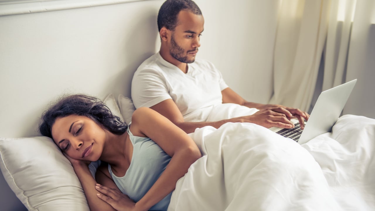 Handsome young Afro American man is using a laptop, lying in bed at home, while his beautiful wife is sleeping near