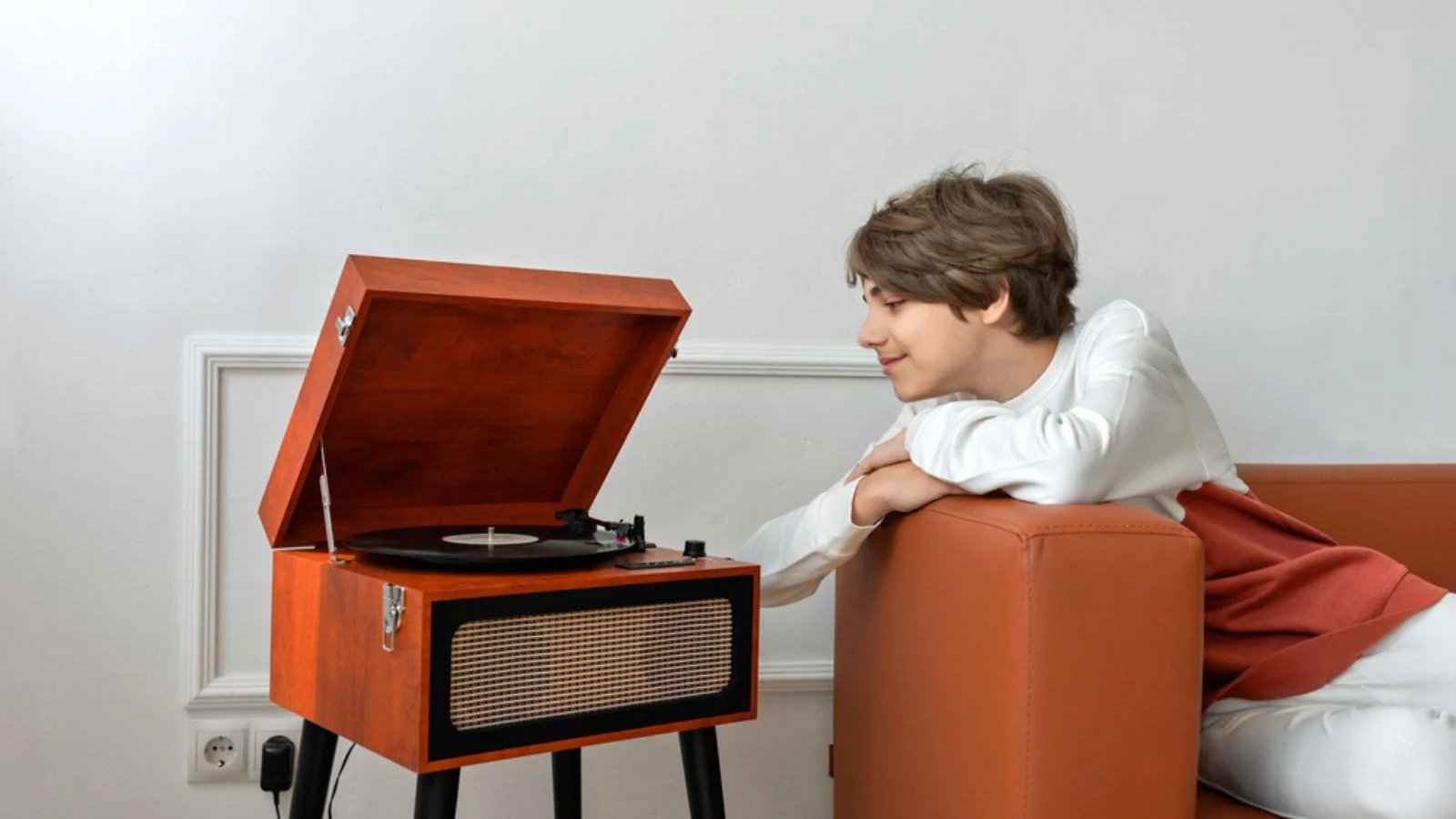 Handsome teenager boy listening vinyl records on a retro wooden turntable, laying on brown sofa and smiling, enjoy music sound