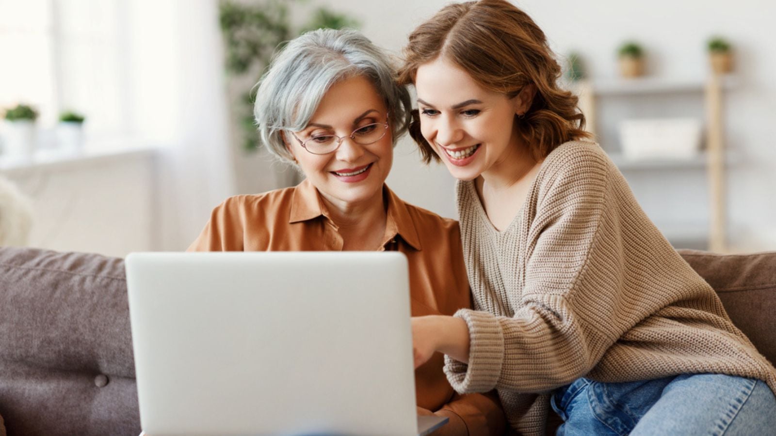 Happy aged woman with young adult daughter smiling while sitting on sofa and using laptop in living room