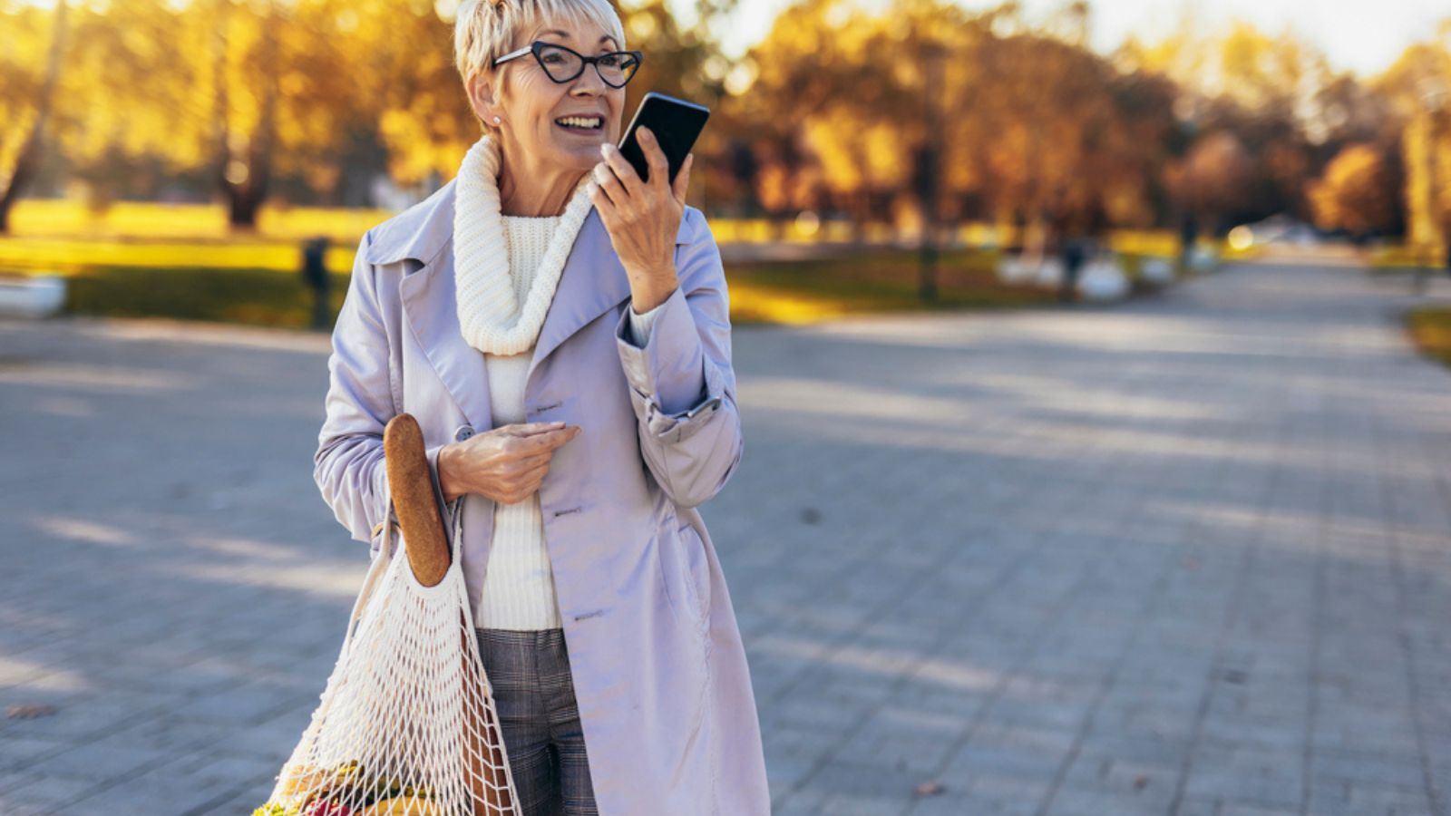 Happy smiling senior woman is holding a reusable bag with fruits and vegetables outdoor using a phone