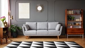 Black and white carpet with geometric pattern placed on the floor in dark living room interior with grey couch, vintage cupboard with books and wainscoting on the wall