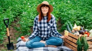Beautiful female farmer meditating while sitting on a blanket near fresh organic vegetables in a wooden box against the background of a vegetable garden