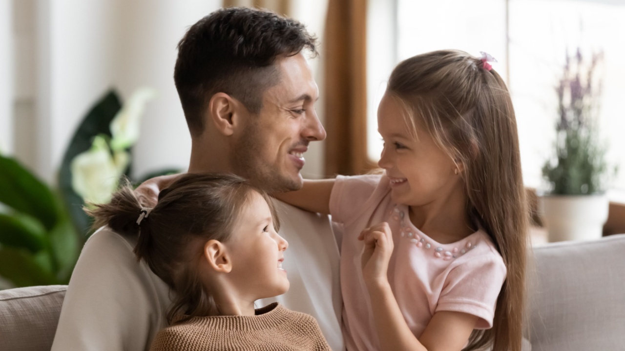 Affectionate cute family young father hugging little preschool children daughters having fun bonding sitting on sofa, smiling dad embracing two small funny kids playing cuddling together at home
