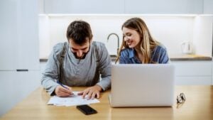 Young bearded caucasian smiling man sitting at dining table and filling in electricity bill. Woman sitting next to him. On table is laptop.