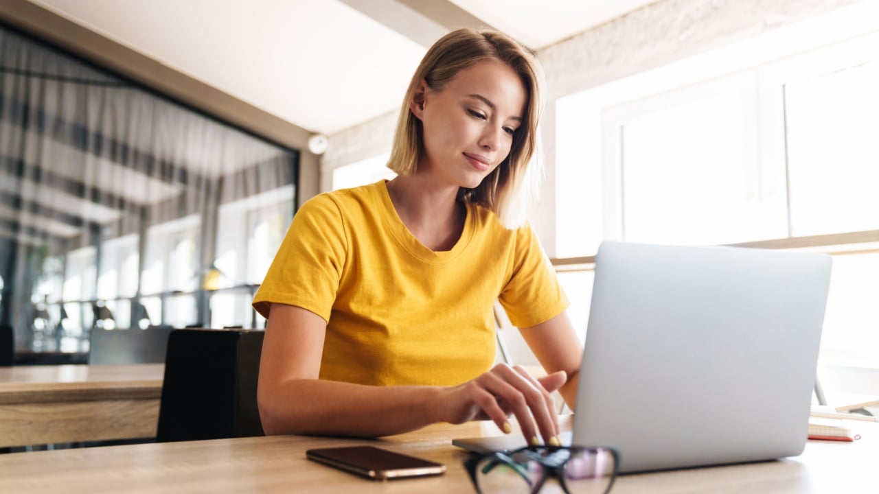 Photo of pleased blonde woman using laptop while sitting at table in open-plan office