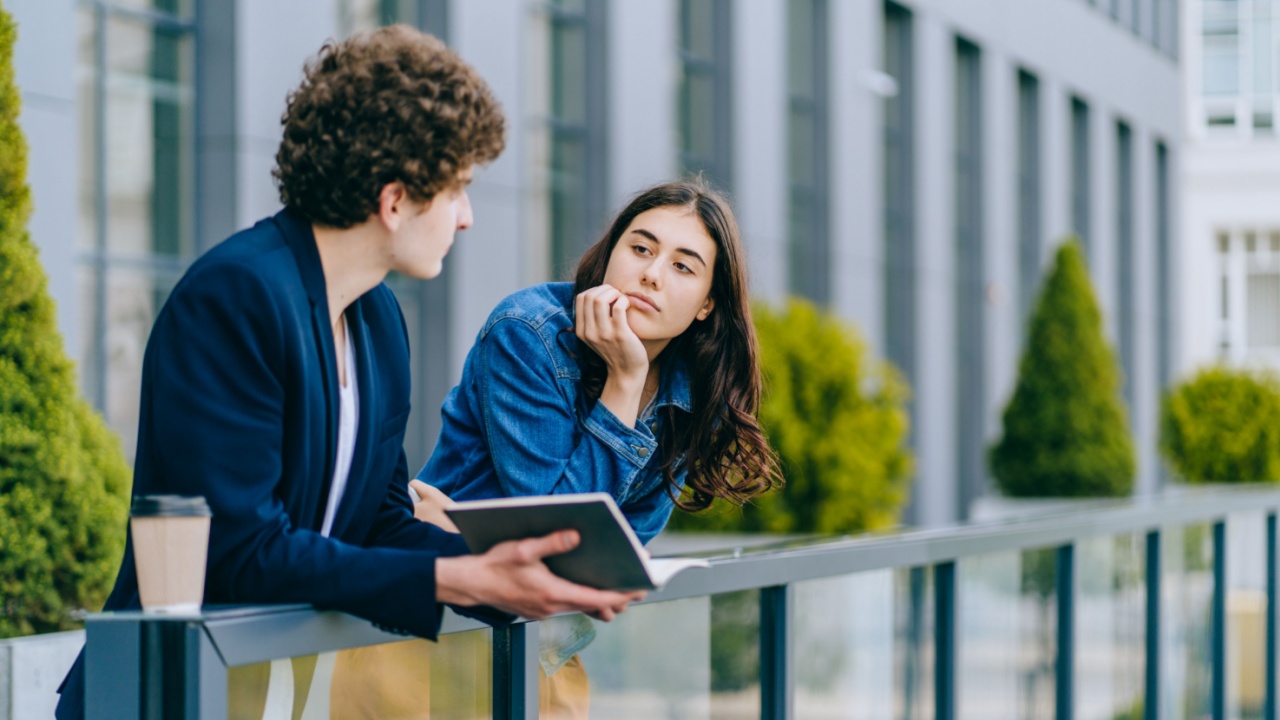 Portrait of charming woman looking aside and boring while man trying to impress with modern urban building on background. Teenagers students on campus of university.