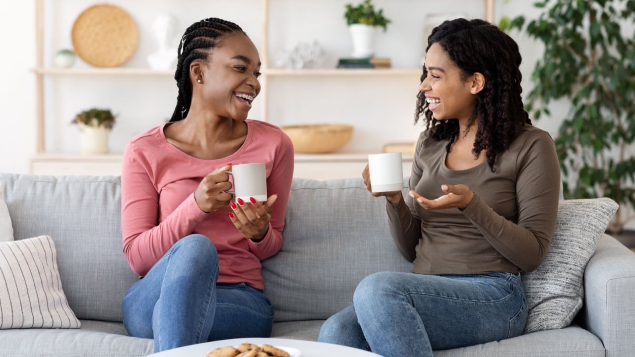 African american girlfriends enjoying weekend together, drinking coffee and talking on couch at home