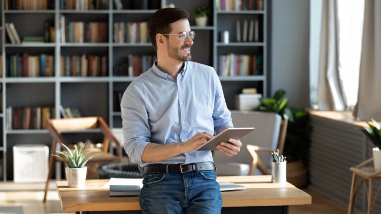 Entrepreneur standing in modern luxury office room distracted from work, business task and planning holding tablet device looks out the window feels happy