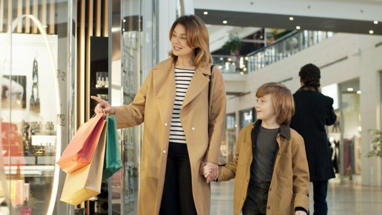 Caucasian mother and small son stepping hand in hand by shopping mall passage and talking. Stop at shop showcase. Good looking woman and boy spending weekend together and looking in store window.