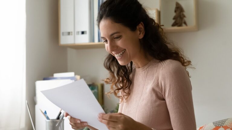 Close up smiling woman reading letter, holding paper sheet, working with correspondence, happy businesswoman or student received good news, money refund or great exam results, domestic bills