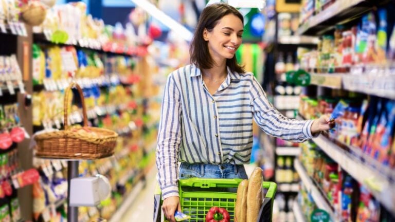 Everyday Shopping Concept. Smiling Young Housewife Pushing Trolley Cart Between Aisles In Grocery Store. Positive Woman Buying Food, Taking Products From Shelf.