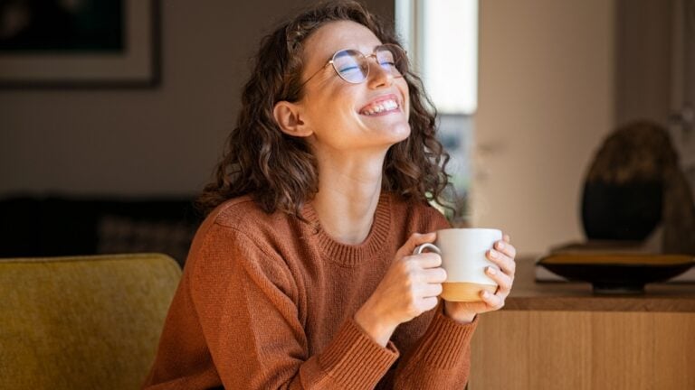 Portrait of joyful young woman enjoying a cup of coffee at home. Smiling pretty girl drinking hot tea in winter. Excited woman wearing spectacles and sweater and laughing in an autumn day.