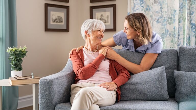 Smiling adult daughter embracing from behind old mother sitting on sofa and laughing together. Aged woman having fun with mature daughter during home visit. Old mom feeling happy with daughter.