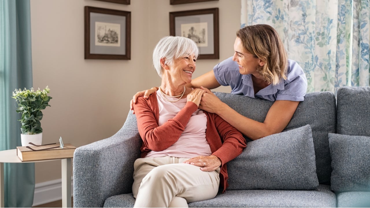 Smiling adult daughter embracing from behind old mother sitting on sofa and laughing together. Aged woman having fun with mature daughter during home visit. Old mom feeling happy with daughter.