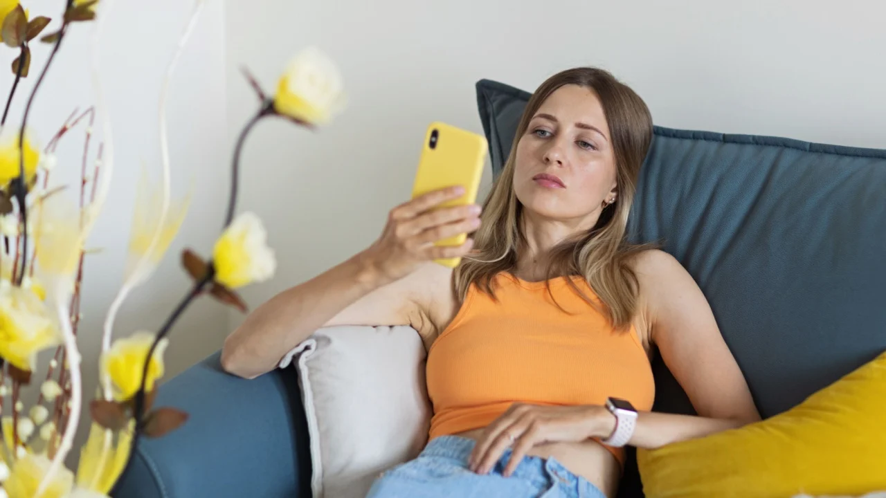 Unhappy young caucasian blonde woman relaxing at home alone, sitting on couch in comfortable pose, blogger sharing good news at social media via mobile phone. Smiling woman enjoying weekend order
