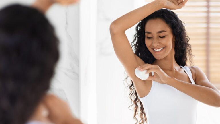 Beautiful Young Woman Applying Roller Deodorant To Armpit Zone In Bathroom, Smiling Millennial Female Using Antiperspirant For Underarms For Sweat Protection, Making Daily Beauty Routine Near Mirror