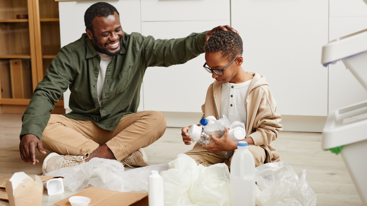 Full length portrait of happy African-American father and son sorting plastic and paper at home for waste recycling.
