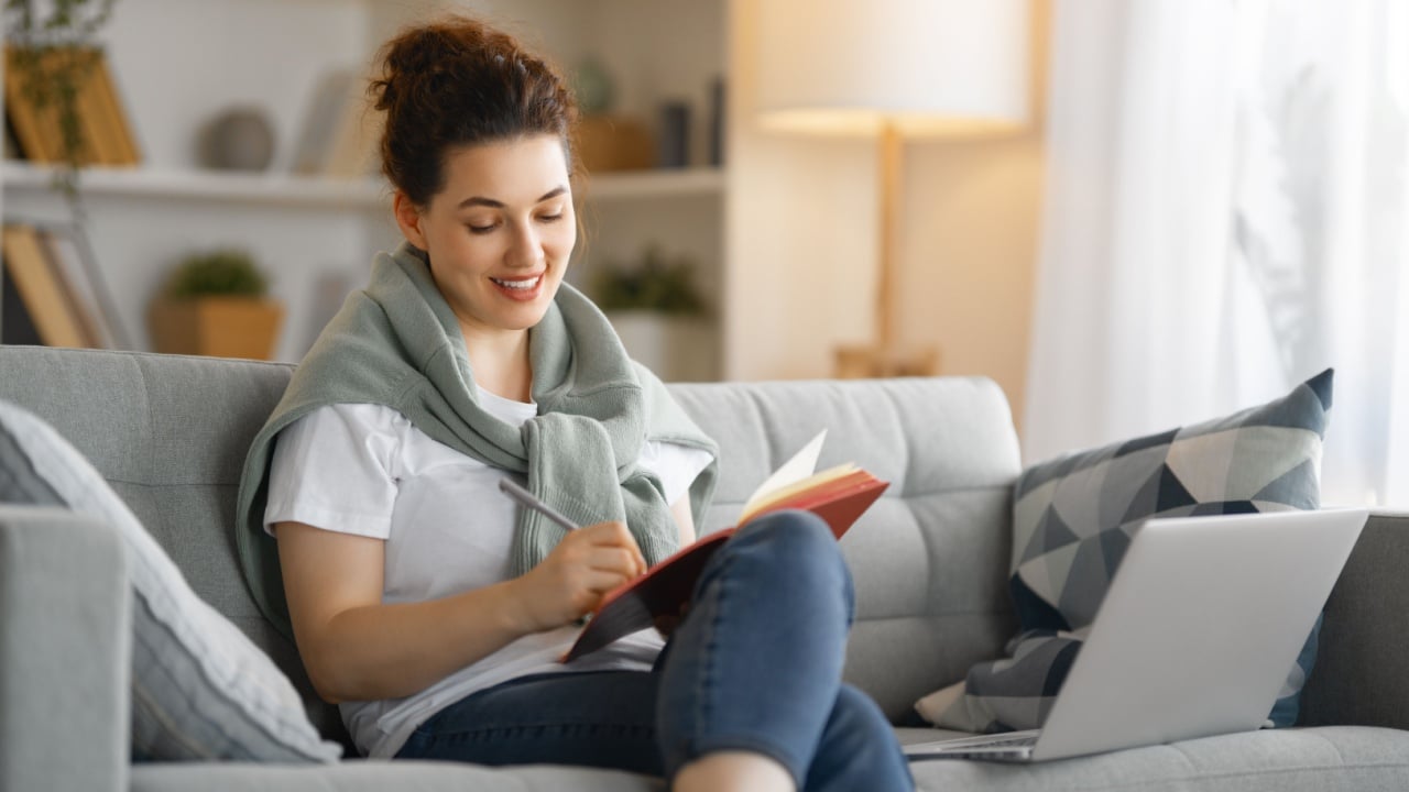 Happy woman studying online, watching webinar, podcast on laptop, making notes.