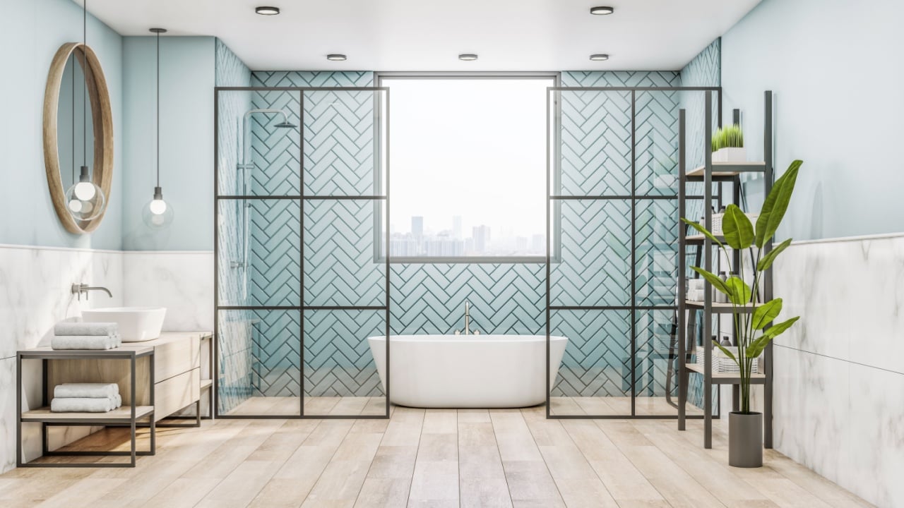 Front view on stylish sunny bathroom with marble walls, green plants wooden floor, wooden round mirror, eco sink cabinet and precious white bath with behind glass partitions.
