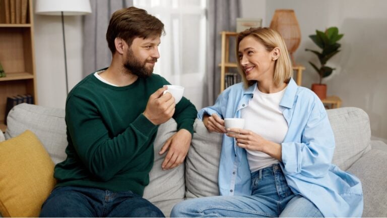 Happy Couple Drinking Coffee At Home. Young Couple Talking And Holding Cups While Sitting On Beige Sofa At Home.