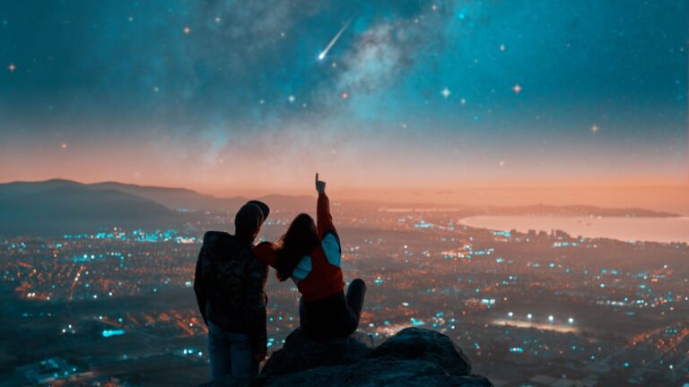 silhouettes of a couple sitting on the top of the mountain looking and pointing out at shooting star and milky way over the city lights on the horizon