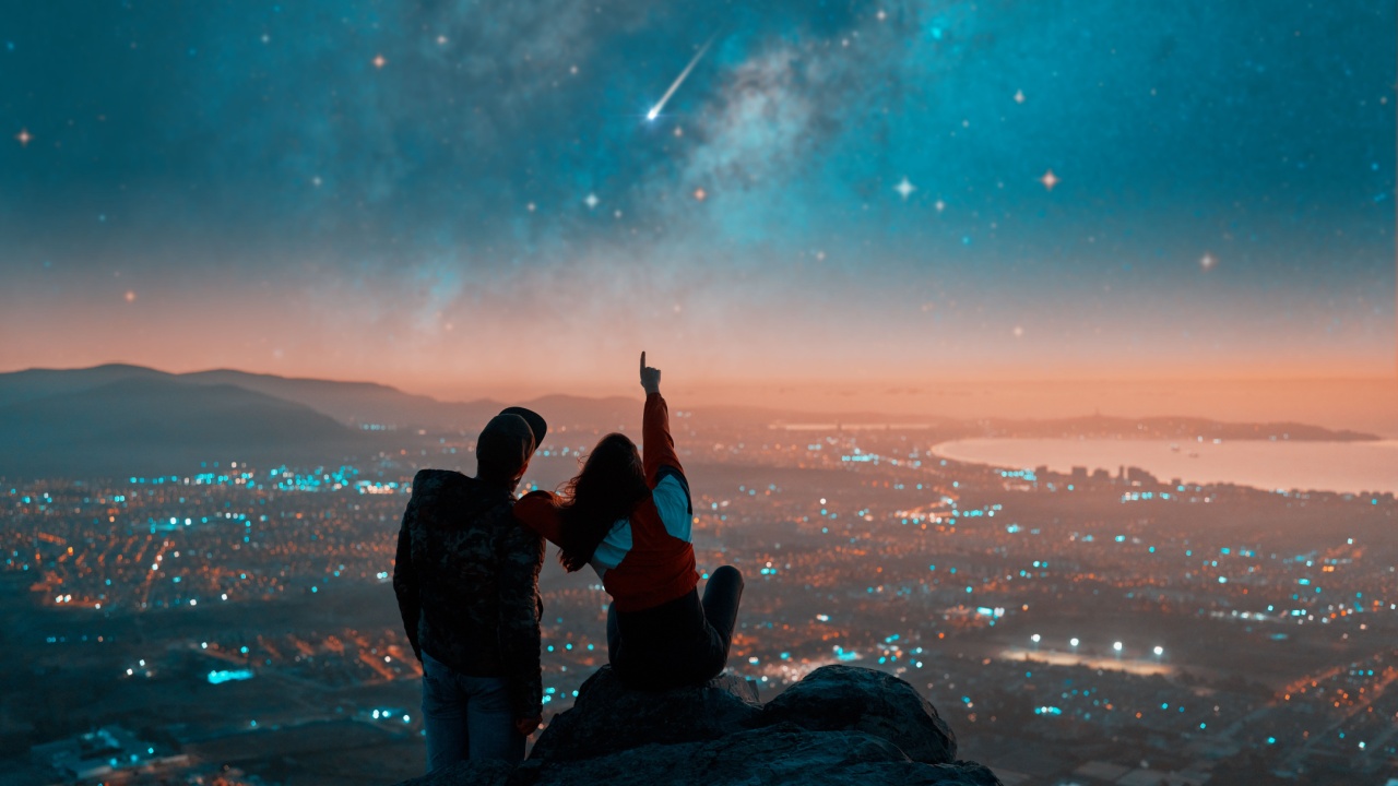 silhouettes of a couple sitting on the top of the mountain looking and pointing out at shooting star and milky way over the city lights on the horizon