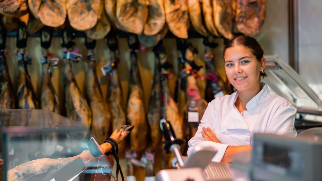 Smiling successful young female butcher shopkeeper standing indoors with crossed arms on background of hanging dry-cured legs of Iberian ham