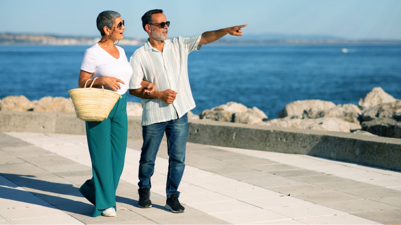 Retirement Travel. Mature Tourists Couple Pointing Finger Aside Looking At Sea View Outdoors, Walking Along Coastline And Enjoying Dream Vacation And Active Retired Life In Summer. Full Length