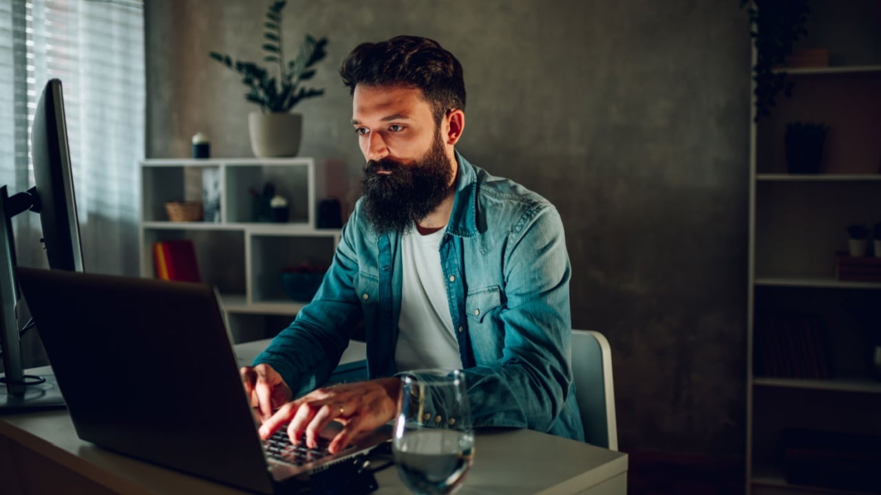 A senior full-stack developer is sitting in his darkened modern cozy home office and typing computer code on his laptop. A full-stack programmer is programming software at his home.