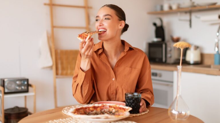 Slice of happiness. Happy young hungry caucasian lady sitting at table in kitchen and eating tasty homemade pizza, enjoying and smiling,