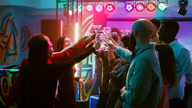 Smiling people clinking glasses filled with alcohol on dance floor, saying cheers under colorful spotlights. Young men and women enjoying cool funky party, entertainment. Handheld shot.