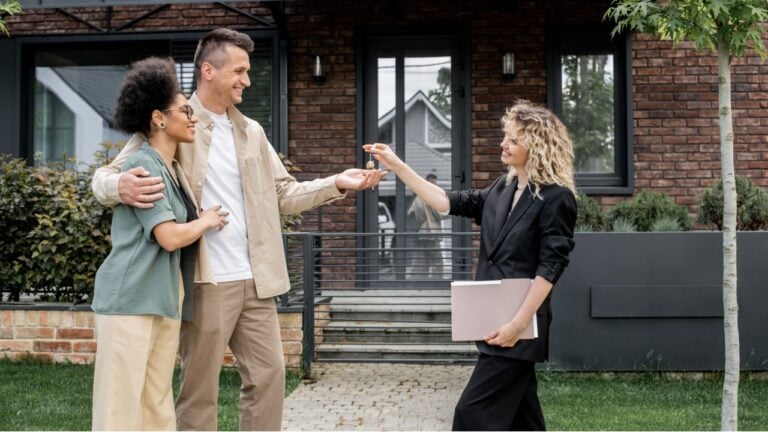 real estate broker with folder giving key to overjoyed interracial couple near new city cottage
