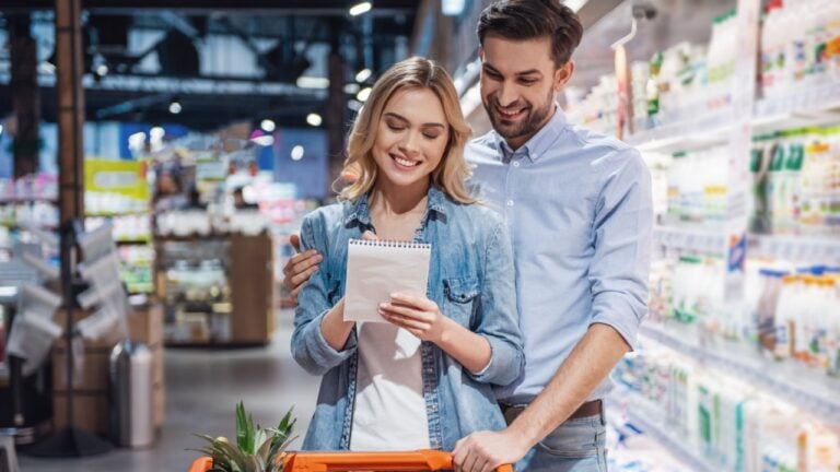 Beautiful couple is reading lists of goods and smiling while doing shopping at the supermarket