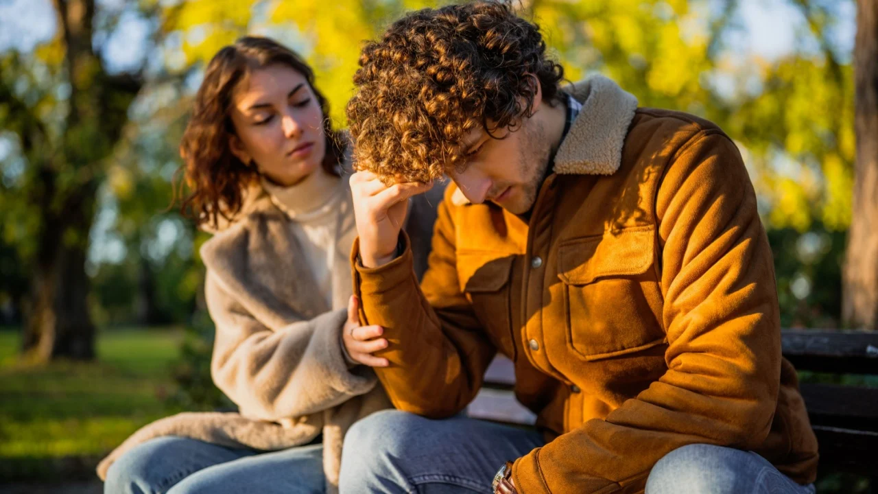 Young couple is sitting in park on sunny day. Man is sad and woman in consoling him.