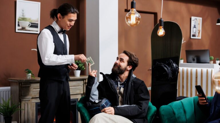 In hotel reception, friendly waiter caters to couple with skiing and snowboarding equipment. While woman is browsing on her cell phone, young man gives money tip to employee in luxury winter resort.