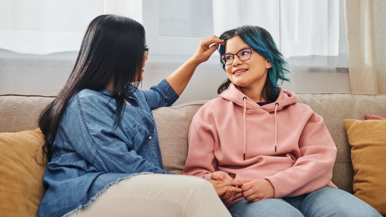 An Asian mother sitting on a couch, tenderly stroking her teenage daughter hair in a cozy home setting.