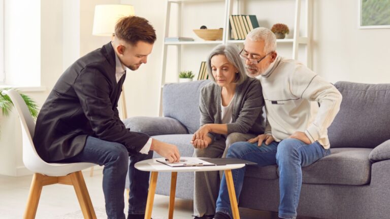 Elderly couple consults with advisor about investments or real estate planning in comfort of home. Young male professional is discussing documents with attentive mature couple in cozy living room.