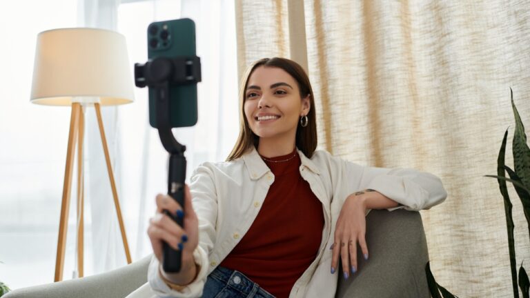 A young woman in casual attire films video content for social media while sitting on a chair in her home office.
