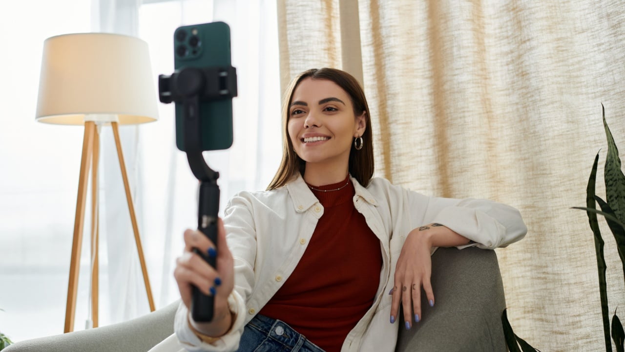 A young woman in casual attire films video content for social media while sitting on a chair in her home office.