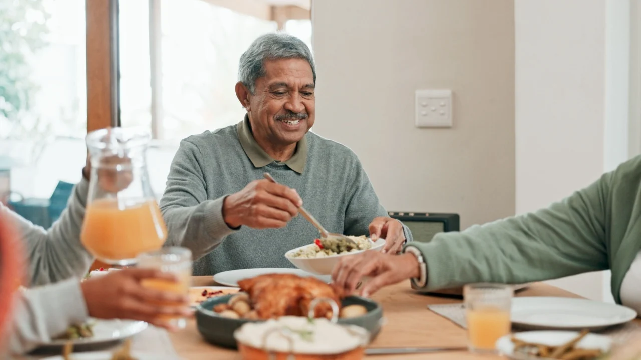Senior man, family and lunch with smile, sharing and helping hand for food, care and connection in home. People, eating and happy with meal on table for reunion, holiday or celebration in dining room