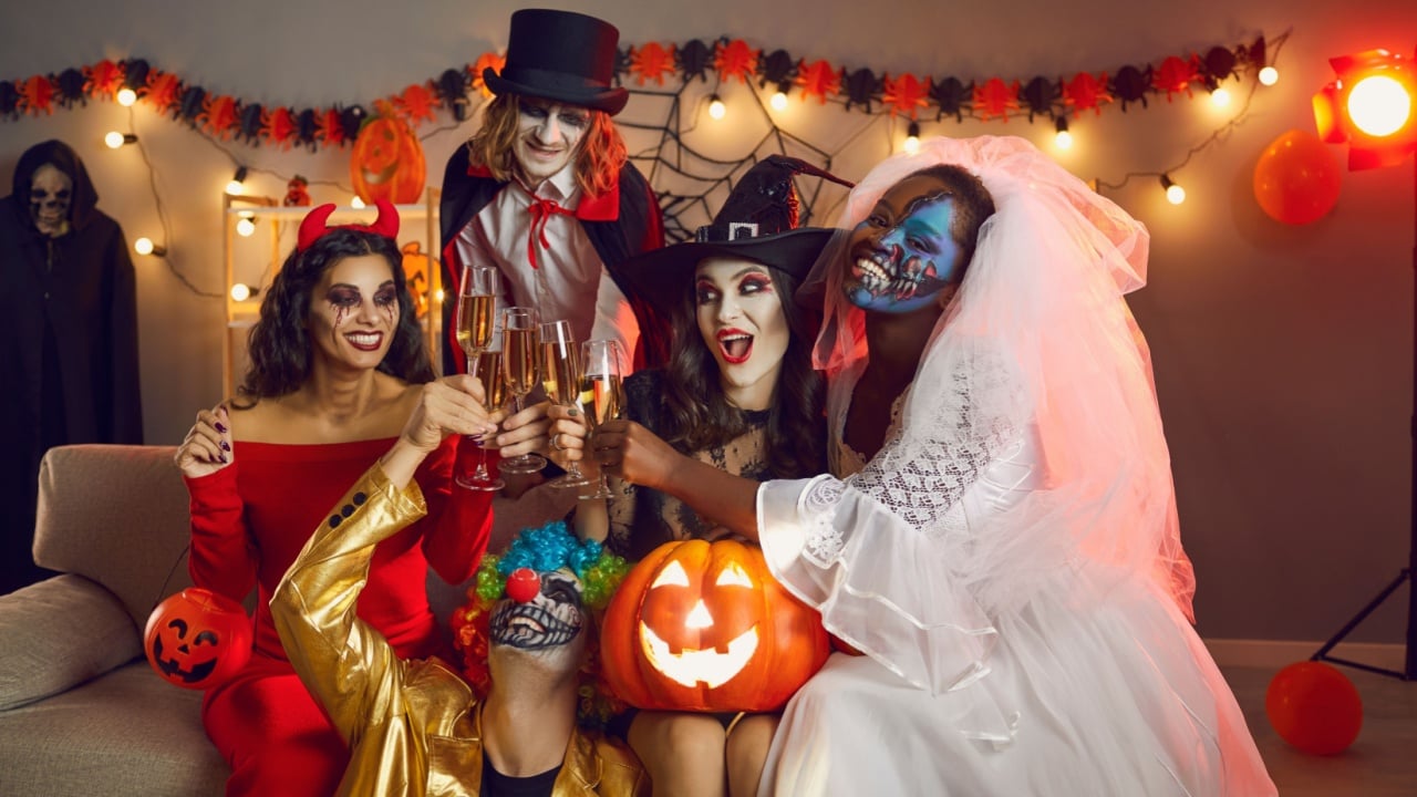 Adult party for Halloween. Group of happy and joyful creative friends having fun clinking glasses of champagne at Halloween party. Multiracial people in various masquerade costumes sit on sofa in room