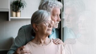 Pensive elderly mature senior man looking in distance out of window, thinking of personal problems. Old bearded male hugging sad wife close to him.